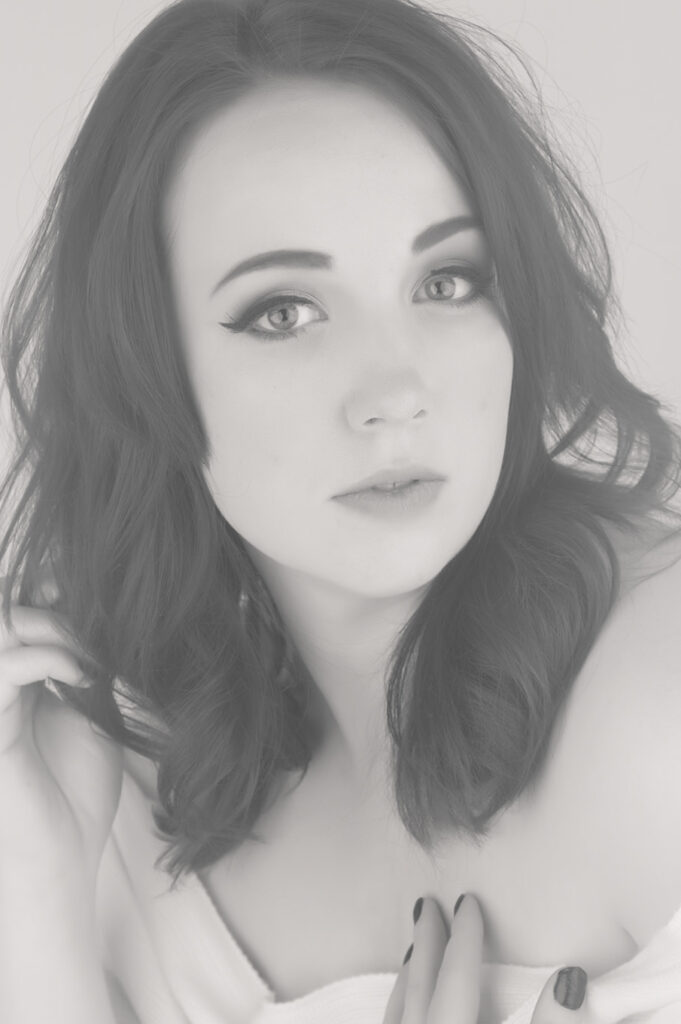 A young woman with wavy dark hair and light eyes gazes softly at the camera during a photography session. Wrapped in white fabric, she poses gracefully in this black and white portrait by a Photographer in Calgary. - Jodi O Photography