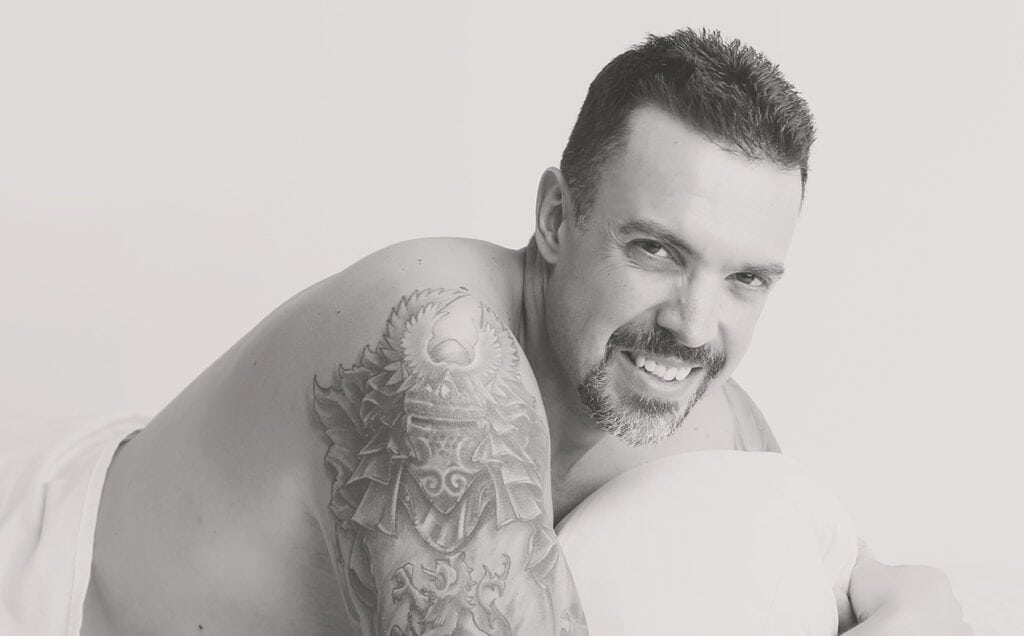 A smiling man with short dark hair and a goatee lies on his side, resting on a pillow. Showcasing Men’s Boudoir in Calgary, he displays a detailed upper arm tattoo in this striking black and white boudoir photography image. - Jodi O Photography