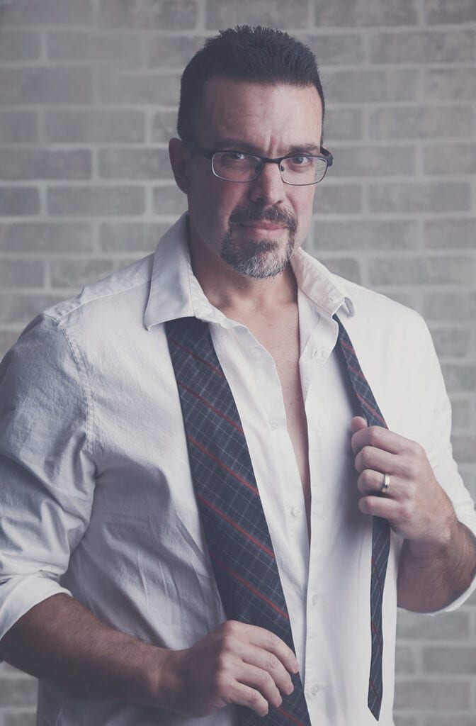 A man with short dark hair, glasses, and a goatee wears an open white shirt with a plaid tie hanging loosely around his neck. He stands confidently before a light brick wall, capturing the essence of stylish Calgary men’s boudoir photography. - Jodi O Photography