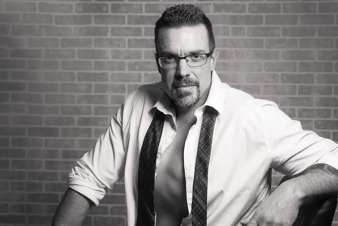 A man with short hair, glasses, and facial hair poses confidently against a brick wall. Embracing men’s boudoir, he wears an open white shirt and a loosely hanging tie, exposing his chest. The image is in black and white. - Jodi O Photography