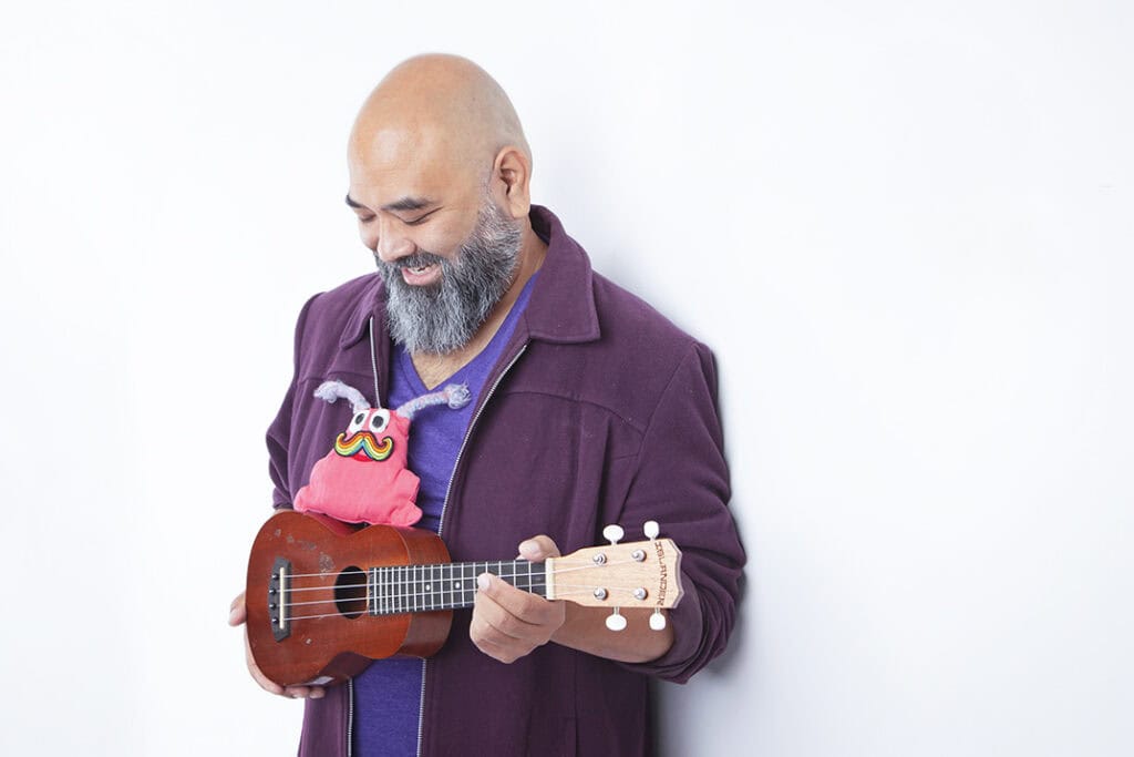 A smiling man with a beard holds a ukulele while looking down. A colorful, plush puppet with googly eyes and antennae sits on his shoulder, bringing Joy to the Stage. He is wearing a purple jacket and stands against a plain white background. - Jodi O Photography