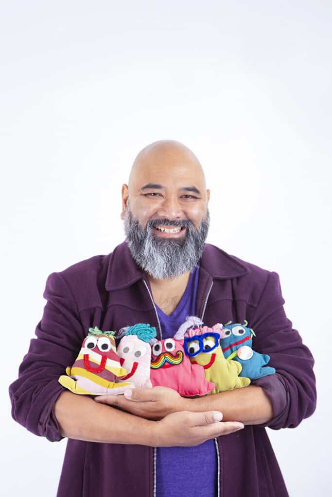 A smiling man with a bald head and gray beard, wearing a purple jacket, brings joy to the stage as he holds five colorful handmade puppets in his arms against a plain white background. - Jodi O Photography