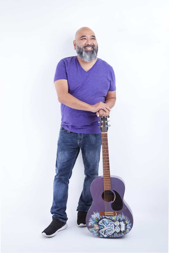 A bearded man wearing a purple shirt and jeans smiles with joy to the stage as he stands, resting his hands on a colorful, decorated acoustic guitar against a plain white background. - Jodi O Photography