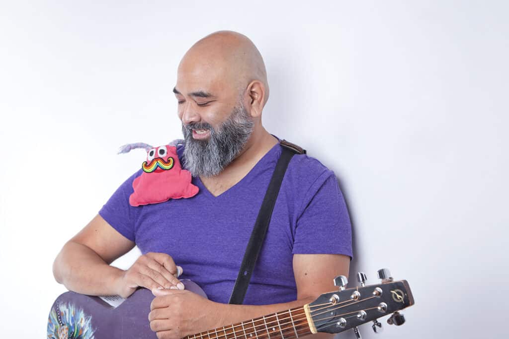 A bearded man wearing a purple shirt smiles while holding a guitar, bringing joy to the stage. A colorful, plush toy with googly eyes and a mustache sits on his shoulder against a plain, white background. - Jodi O Photography