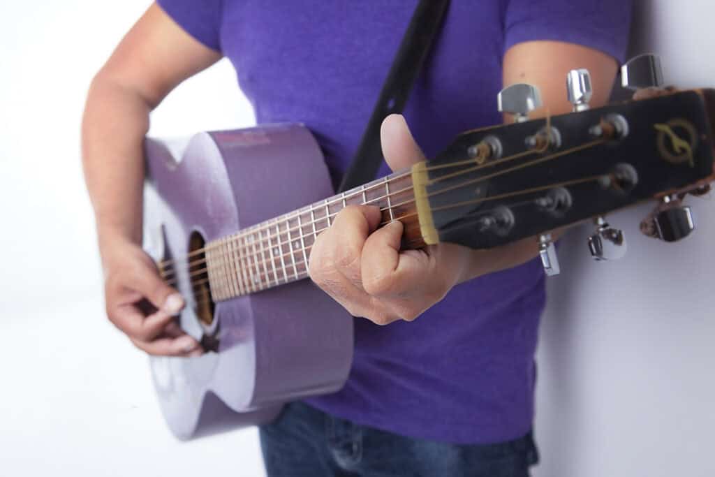 A person wearing a purple shirt brings joy to the stage as they play an acoustic guitar, focusing on their hands strumming and pressing the strings against the fretboard. - Jodi O Photography