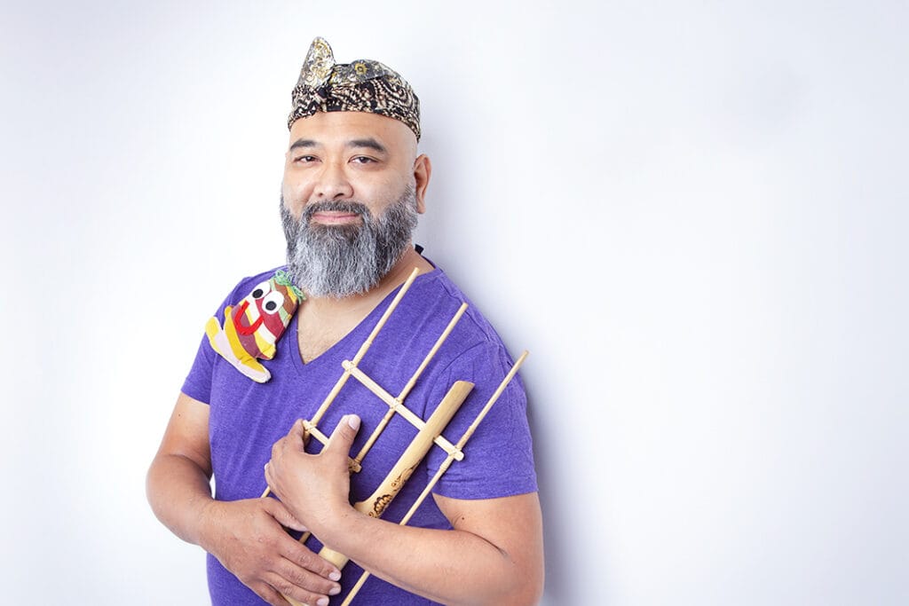 A bearded man wearing a patterned cap and a purple shirt brings joy to the stage, holding bamboo instruments and a colorful cloth puppet on his shoulder, standing against a plain white background. - Jodi O Photography
