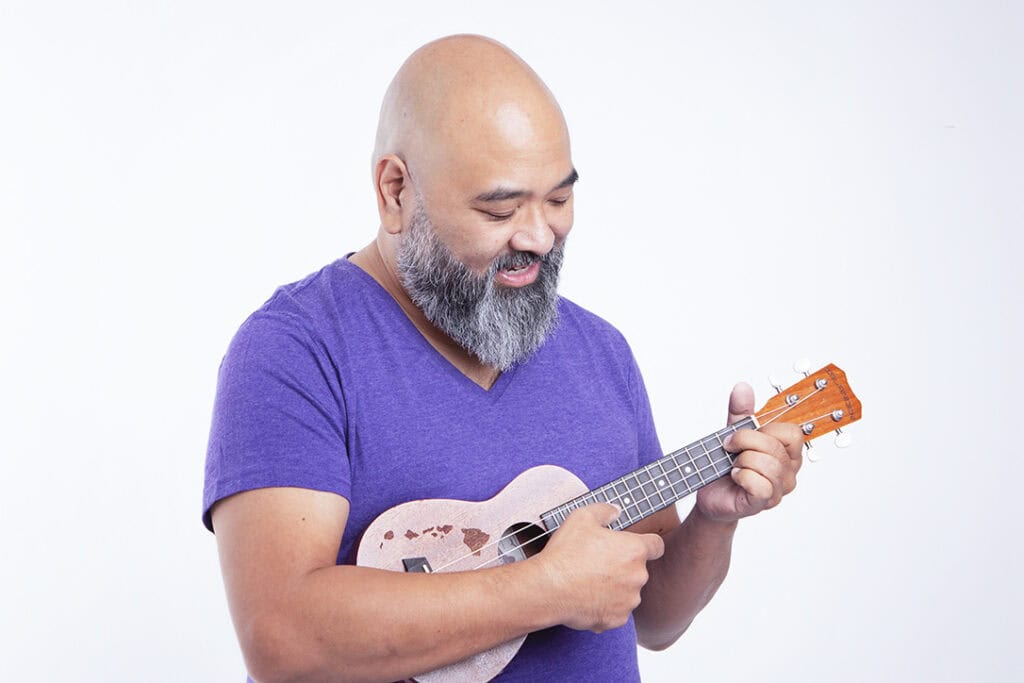 A smiling man with a gray beard and bald head, wearing a purple shirt, strums a small ukulele in a joyful performance against a plain white background. - Jodi O Photography