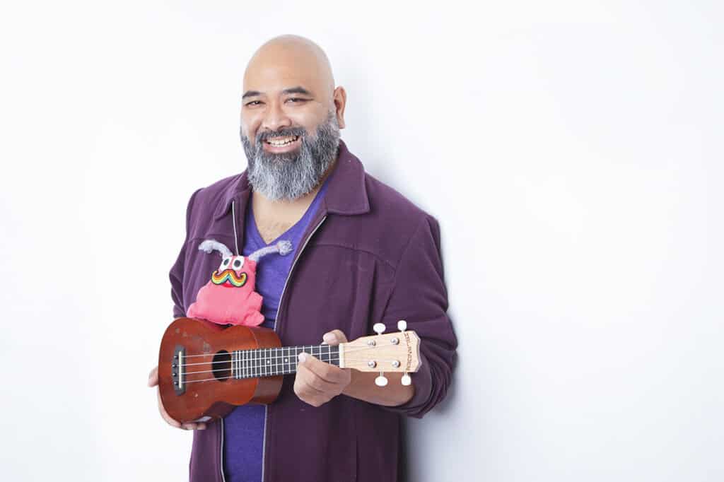 A smiling man with a gray beard holds a ukulele with a colorful, cartoonish puppet sitting on top, bringing joy to the stage. He wears a purple jacket and stands against a plain white background. - Jodi O Photography