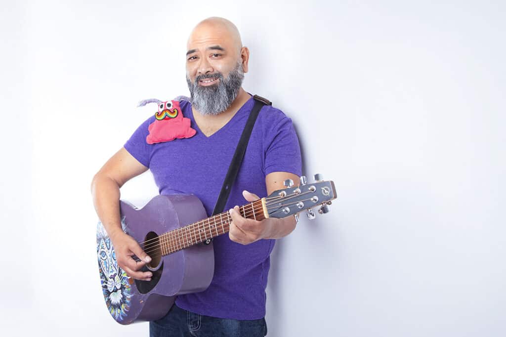 A bearded man in a purple shirt brings joy to the stage as he plays a purple acoustic guitar, smiling gently. A pink plush toy with eyes and a mustache sits on his shoulder, set against a plain white background. - Jodi O Photography
