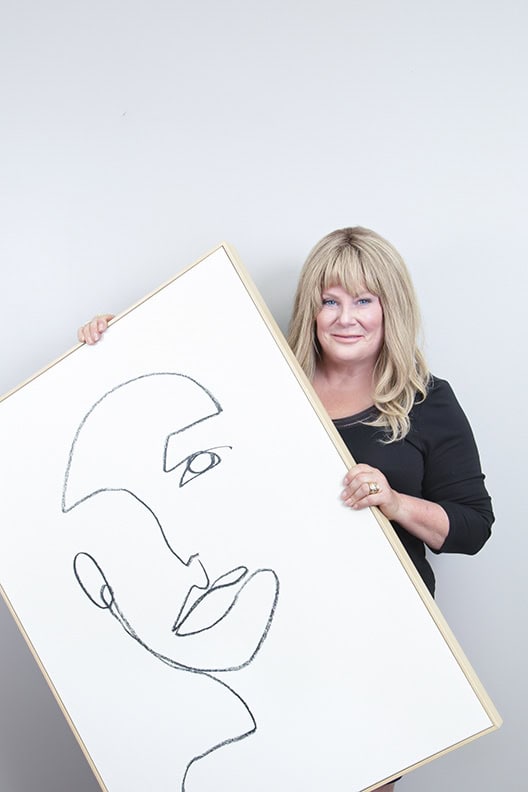 A woman with blonde hair stands against a plain light wall, smiling with confidence and holding a large framed minimalist line drawing of an abstract face. - Jodi O Photography