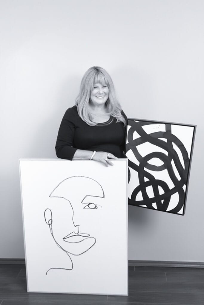 A woman with long, light-colored hair stands smiling with confidence, holding two framed abstract artworks—one features a minimalist line drawing of a face, and the other displays bold black curved lines. - Jodi O Photography