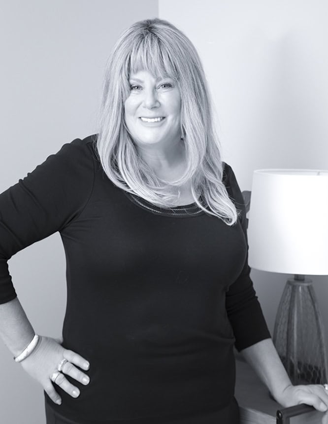 A woman with long, light hair wearing a black top stands indoors, smiling at the camera. With sass and confidence, one hand rests on her hip and the other on a table beside a lamp. The image is in black and white. - Jodi O Photography