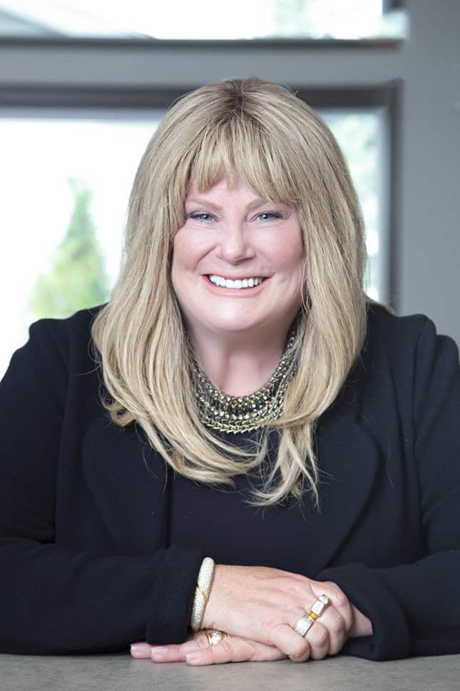 A woman with long blonde hair, exuding confidence and style in a black blazer and layered necklace, smiles while resting her arms on a table. She is indoors with a large window and blurred greenery in the background. - Jodi O Photography