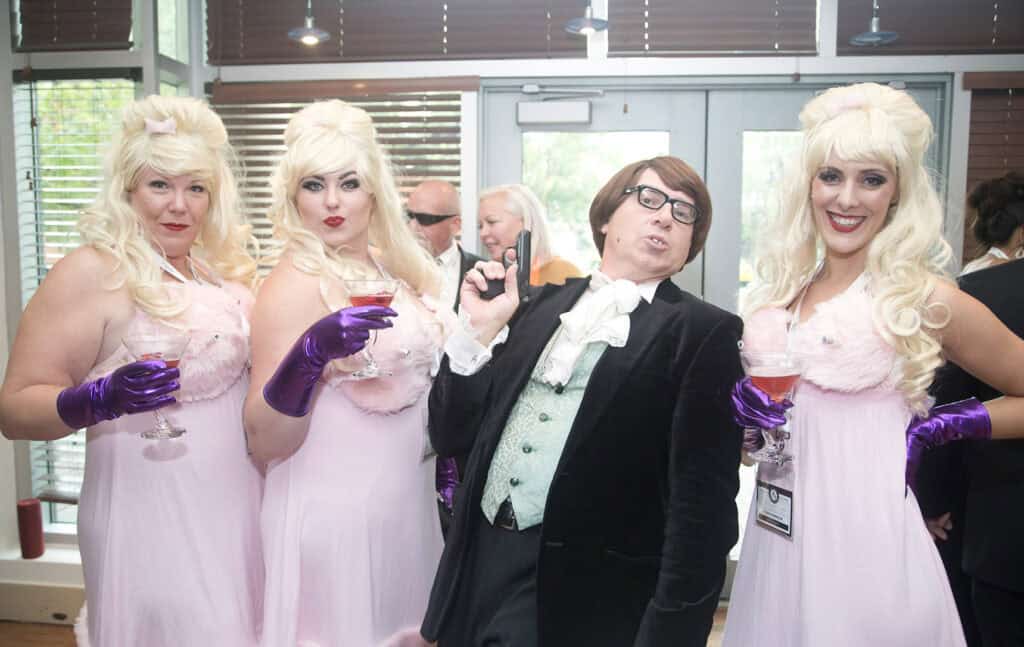 Three people in matching pink dresses, blonde wigs, and purple gloves pose with someone in a black suit at a lively indoor event—perfectly captured by a Calgary Corporate Event Photographer experienced in business event photography. - Jodi O Photography
