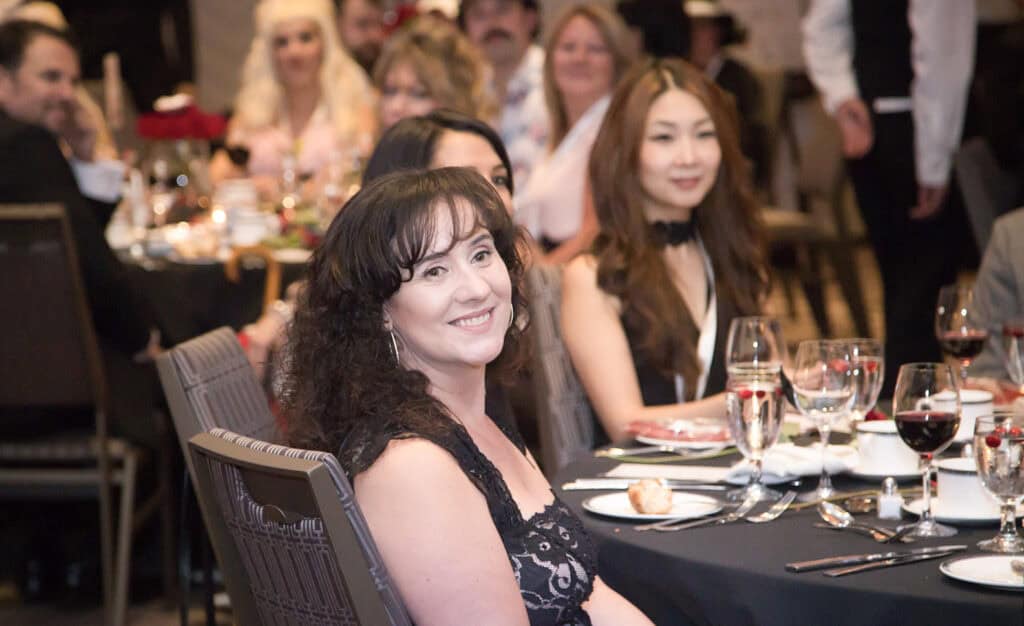 A woman in a black lace dress smiles while seated at a formal business event. Other guests, elegantly dressed, sit at round tables decorated with red and white flowers and glassware—captured by a Calgary Corporate Event Photographer. - Jodi O Photography