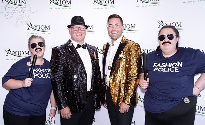 Four people pose in front of an Axiom Mortgage Solutions backdrop; two wear sequin jackets and bow ties, while two in "Fashion Police" shirts, sunglasses, and toy batons strike a fun pose—perfect for a Calgary corporate event photographer to capture. - Jodi O Photography