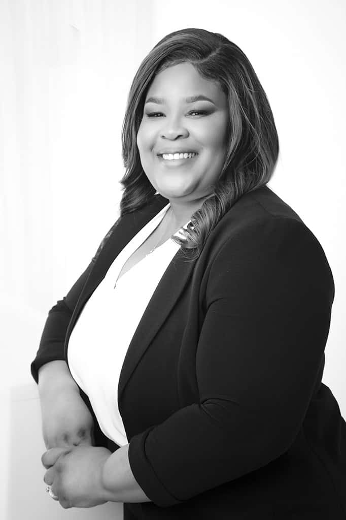 A woman with long, wavy hair smiles warmly while wearing a dark blazer over a light top, posing with her arms resting on a surface against a bright, neutral background. - Jodi O Photography
