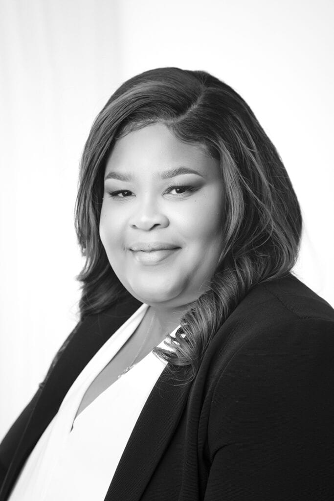 Black and white portrait of a woman with long, wavy hair wearing a dark blazer over a light blouse, smiling softly at the camera against a plain, light background. - Jodi O Photography