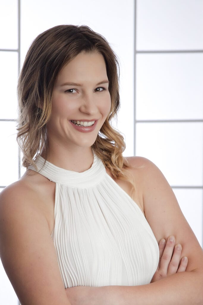 A woman with wavy light brown hair, wearing a sleeveless white top, smiles with arms crossed in front of her against a bright, modern background with rectangular patterns—perfect for business headshots in Calgary. - Jodi O Photography