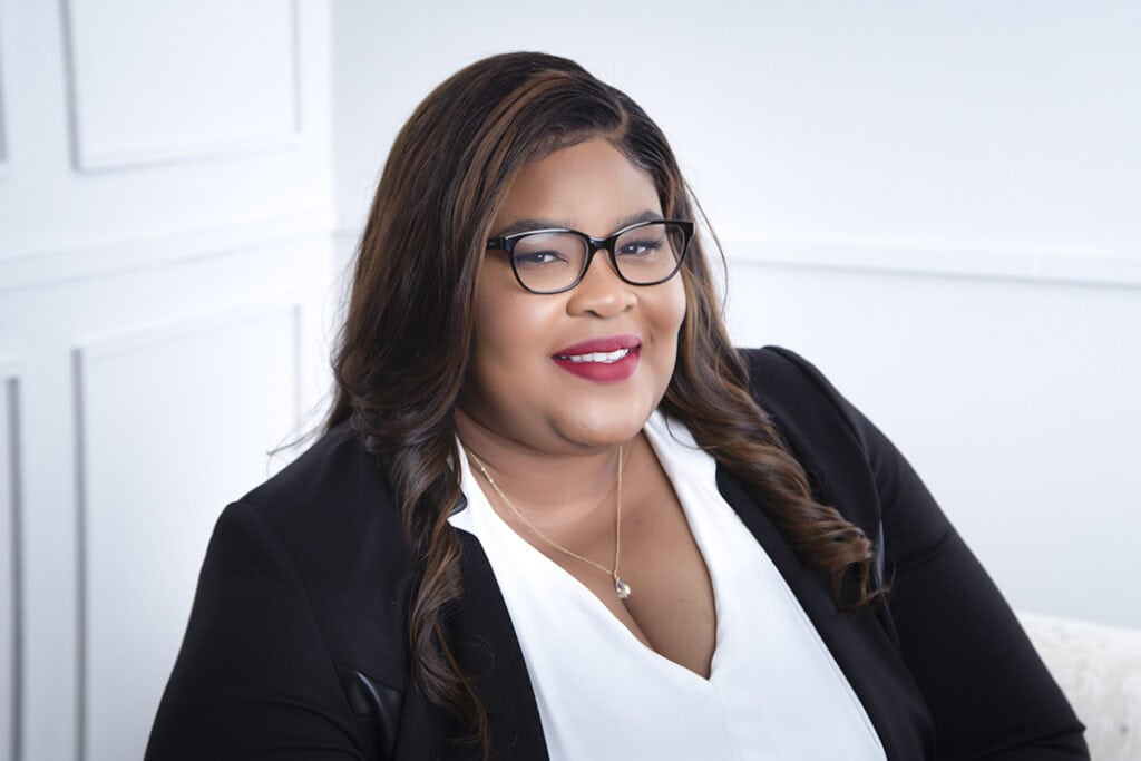 A woman with long wavy hair and glasses smiles at the camera. She is wearing a black blazer over a white blouse and a gold necklace, seated against a light-colored background. - Jodi O Photography