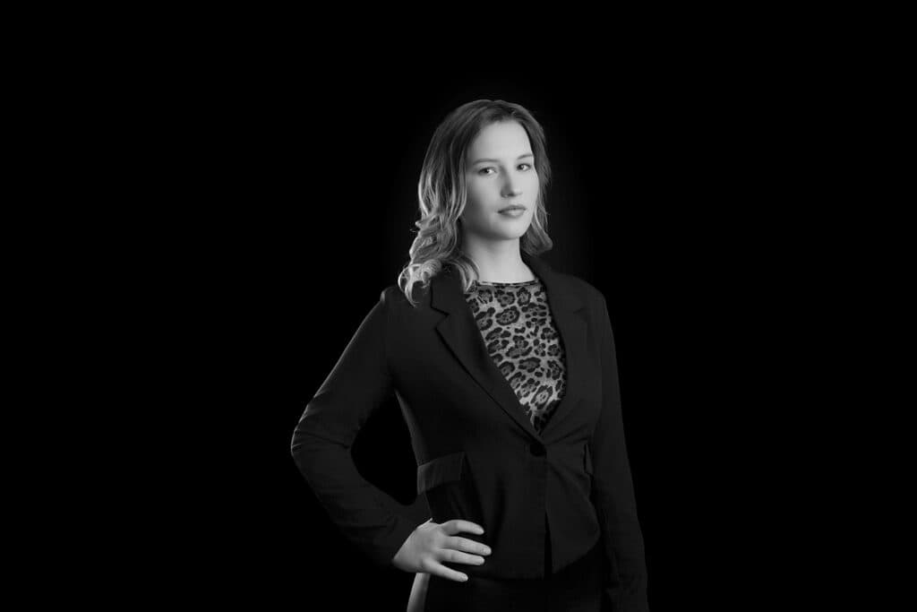 Black and white portrait of a confident woman standing with one hand on her hip, wearing a dark blazer over a leopard print top—perfect for modern business headshots in Calgary—looking directly at the camera against a plain black background. - Jodi O Photography