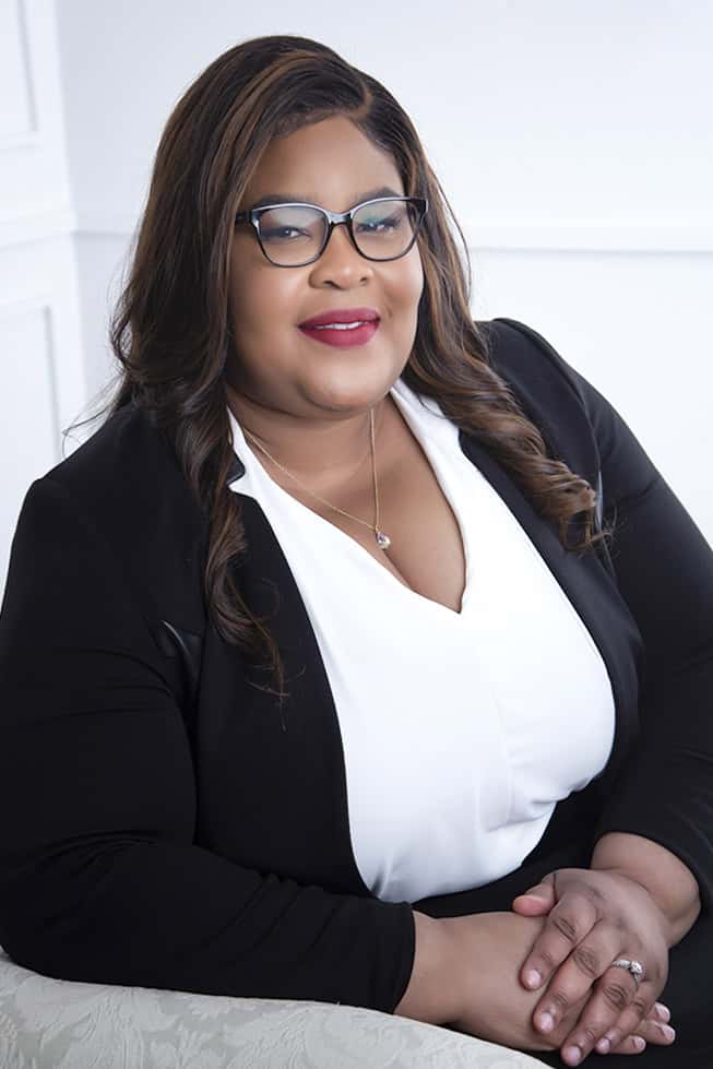 A woman with long brown hair and glasses is smiling while seated, wearing a black blazer over a white blouse. She has a gold necklace and deep red lipstick, and her hands are folded on her lap. The background is light and simple. - Jodi O Photography