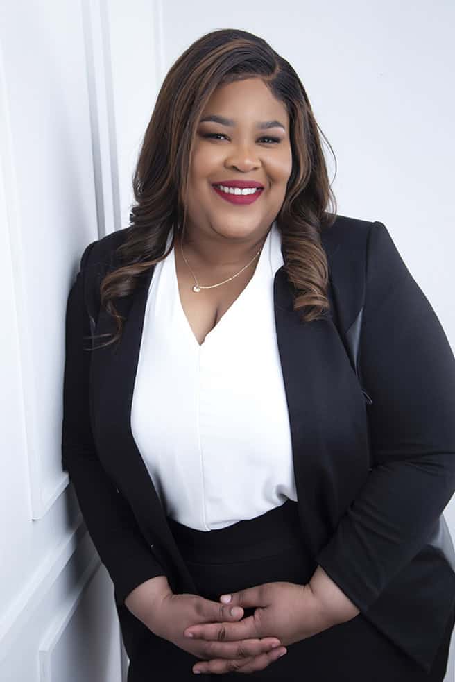 A woman with long, wavy hair wearing a black blazer over a white blouse smiles while standing against a white wall with her hands folded in front of her. - Jodi O Photography