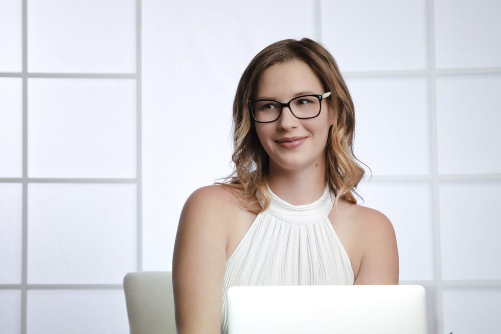 A woman with shoulder-length blonde hair and glasses, wearing a sleeveless white top, sits in front of a laptop and smiles slightly. The bright background with large window panes creates the perfect setting for modern business headshots Calgary professionals love. - Jodi O Photography