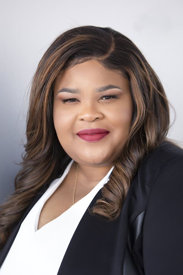 A woman with long, wavy brown hair wearing a black blazer over a white top, smiling softly in a professional headshot against a light gray background. - Jodi O Photography