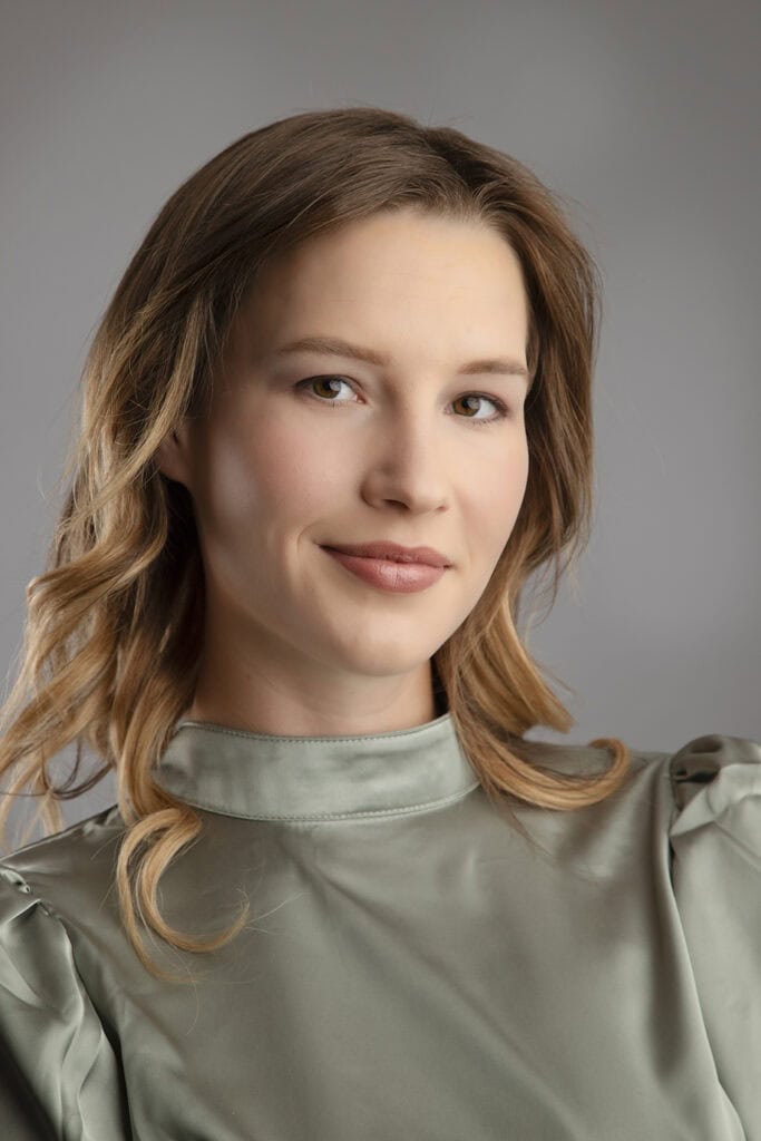 A woman with light brown hair and fair skin, wearing a sage green high-neck blouse, poses against a neutral gray background for Calgary business headshots, smiling softly at the camera. - Jodi O Photography