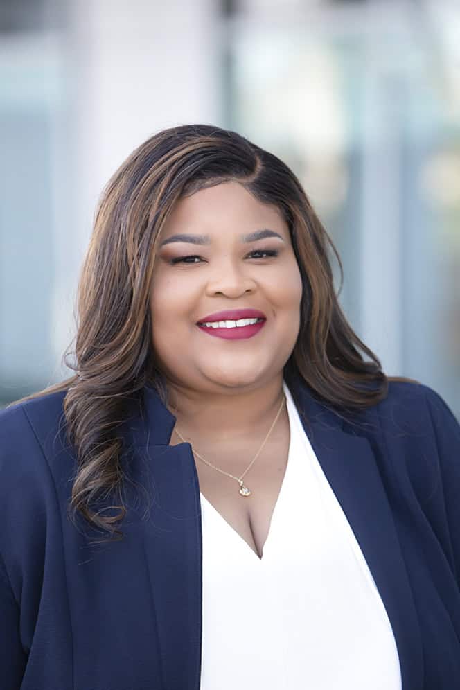 A woman with long, wavy brown hair smiles at the camera. She is wearing a navy blazer over a white top and a gold necklace. The background is softly blurred. - Jodi O Photography