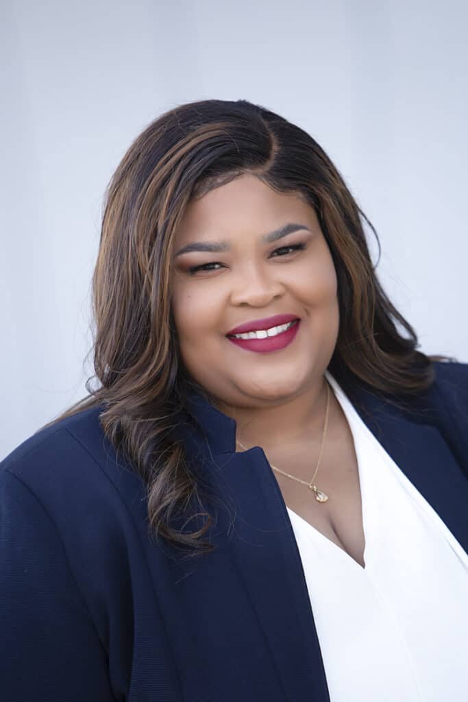 A woman with long, wavy brown hair smiling, wearing a navy blazer over a white top and a gold necklace, standing in front of a plain light-colored background. - Jodi O Photography