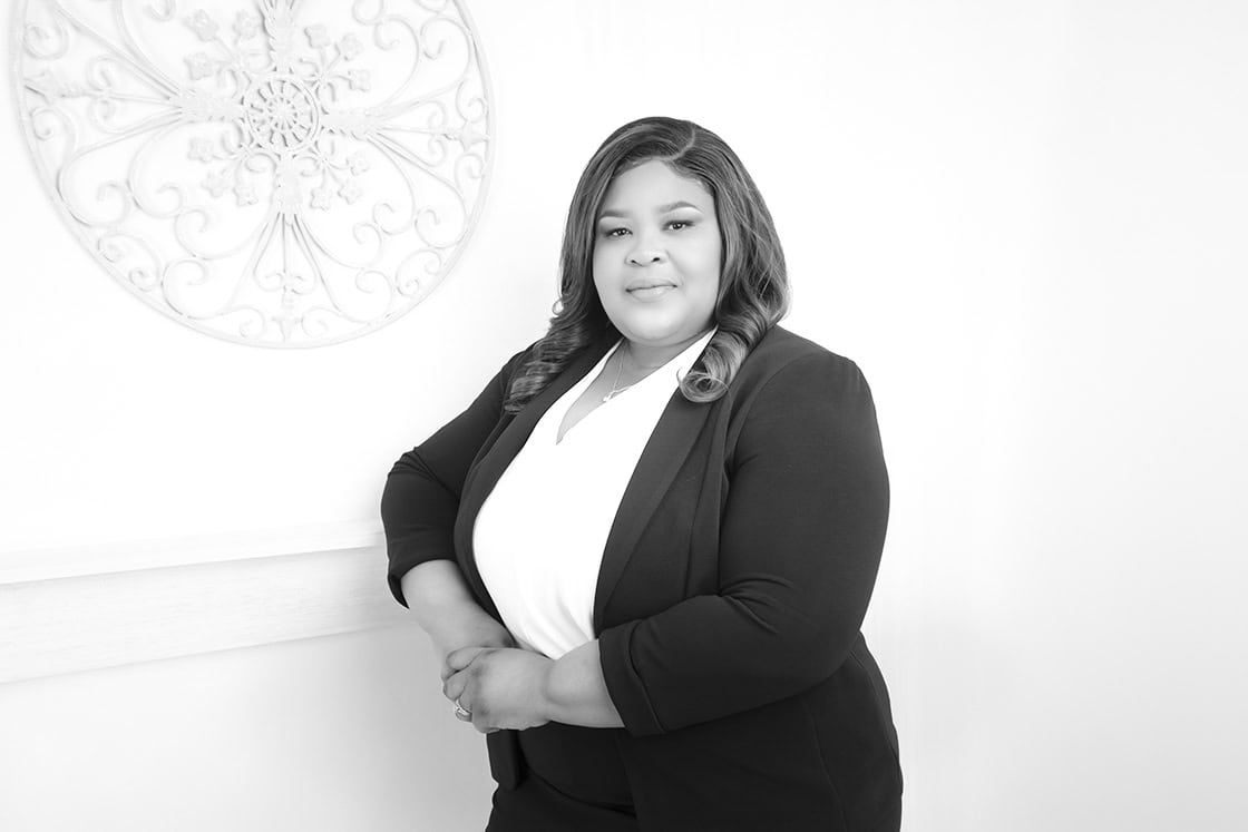 A woman in a dark suit and white top stands indoors, smiling and posing confidently with her hands clasped. There is decorative wall art in the background. The photo is in black and white. - Jodi O Photography