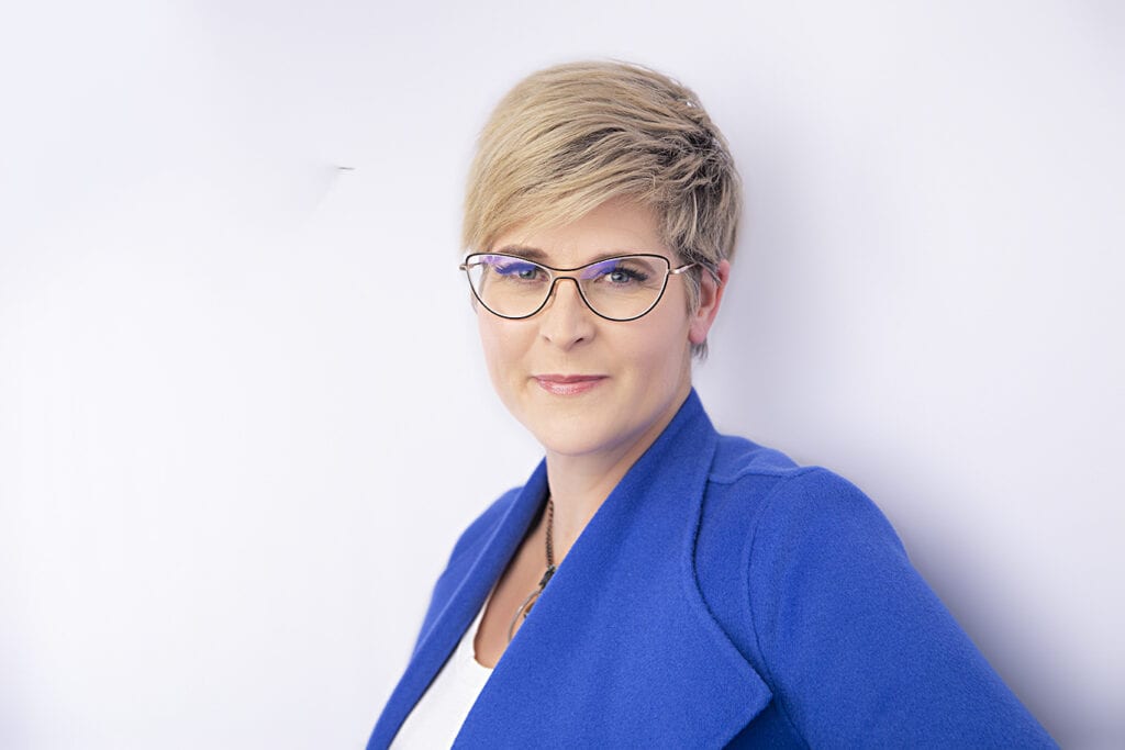 A person with short blonde hair and glasses exudes confidence in a bright blue blazer over a white top, standing against a plain white background and smiling slightly at the camera. - Jodi O Photography