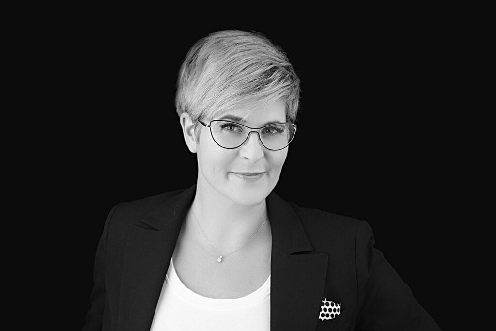 A person with short, light hair and glasses exudes confidence and style in a dark blazer over a white top, smiling slightly at the camera against a solid black background. - Jodi O Photography