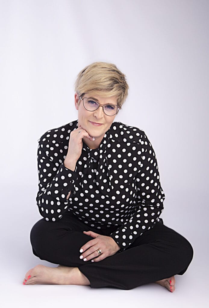 A woman with short blonde hair and glasses exudes confidence and style in a black and white polka dot blouse and black pants, sitting cross-legged on the floor, smiling softly with her hand near her chin against a light background. - Jodi O Photography