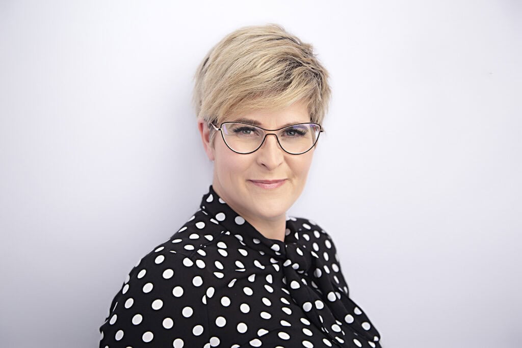 A woman with short blonde hair and glasses wears a black blouse with white polka dots, exuding subtle style as she smiles gently against a plain light background. - Jodi O Photography