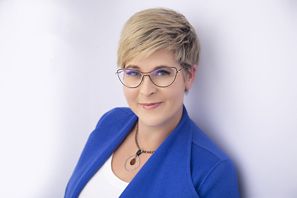 A woman with short blonde hair, wearing glasses, a blue blazer, white top, and a pendant necklace, exudes confidence as she smiles slightly while posed against a plain white background. - Jodi O Photography