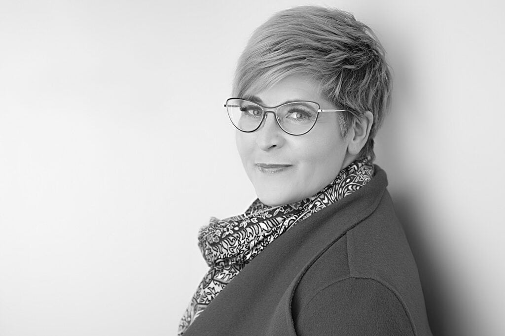 Black and white portrait of a person with short hair and glasses, wearing a patterned scarf and coat, looking at the camera with sass and confidence, a slight smile adding style against a plain background. - Jodi O Photography