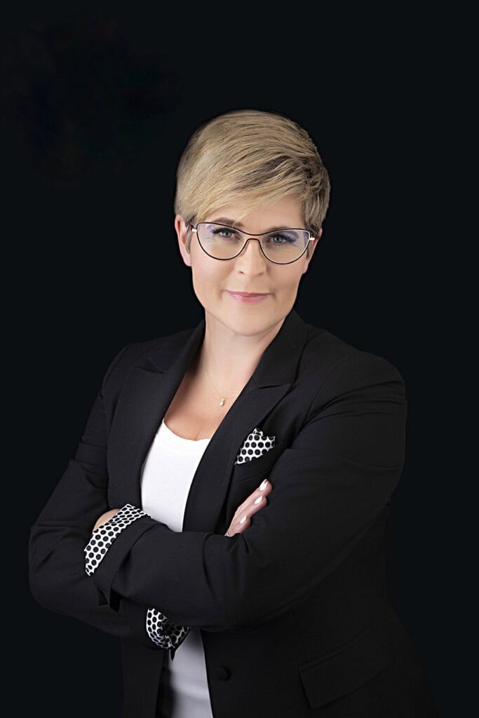 A person exuding confidence and style, with short blonde hair and glasses, stands with arms crossed in a black blazer over a white top against a plain black background. - Jodi O Photography