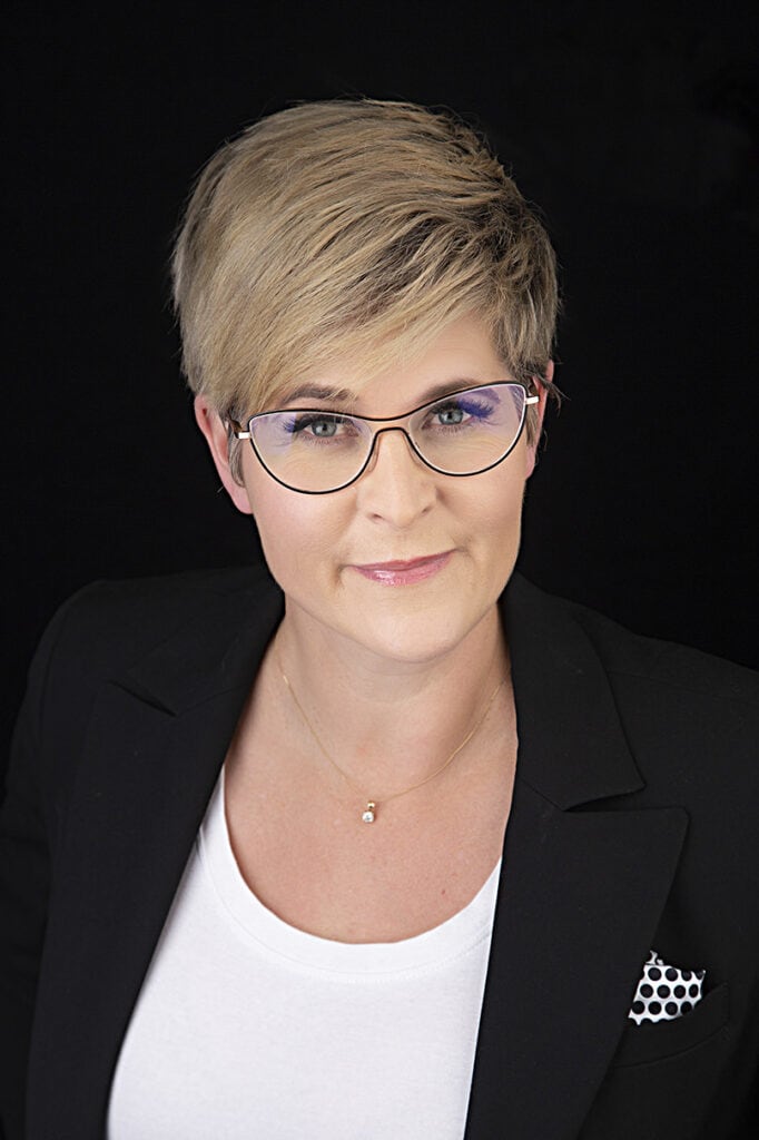 A person with short blonde hair, wearing glasses, a black blazer, and a white top, exudes confidence as they pose against a black background and smile softly at the camera. - Jodi O Photography