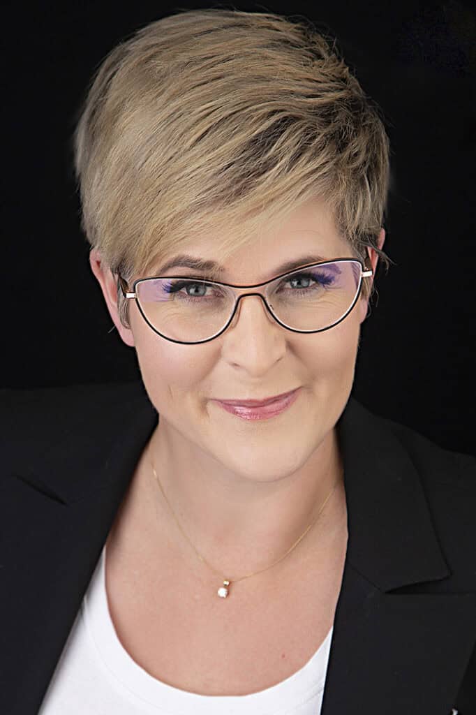 A person with short, light brown hair and rectangular glasses exudes confidence and style in a black blazer and white top, smiling at the camera against a black background. - Jodi O Photography