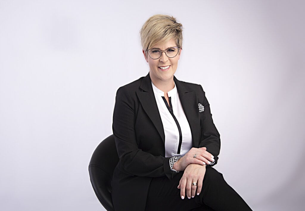 A person with short blonde hair and glasses, dressed in a black suit and white shirt, sits confidently with a hint of sass, smiling on a black chair against a plain light background. - Jodi O Photography