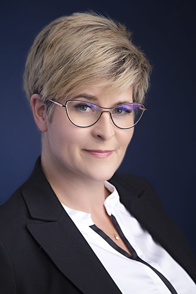 A woman with short blonde hair, glasses, and a black blazer over a white blouse poses against a dark blue background, her slight smile radiating confidence and style. - Jodi O Photography