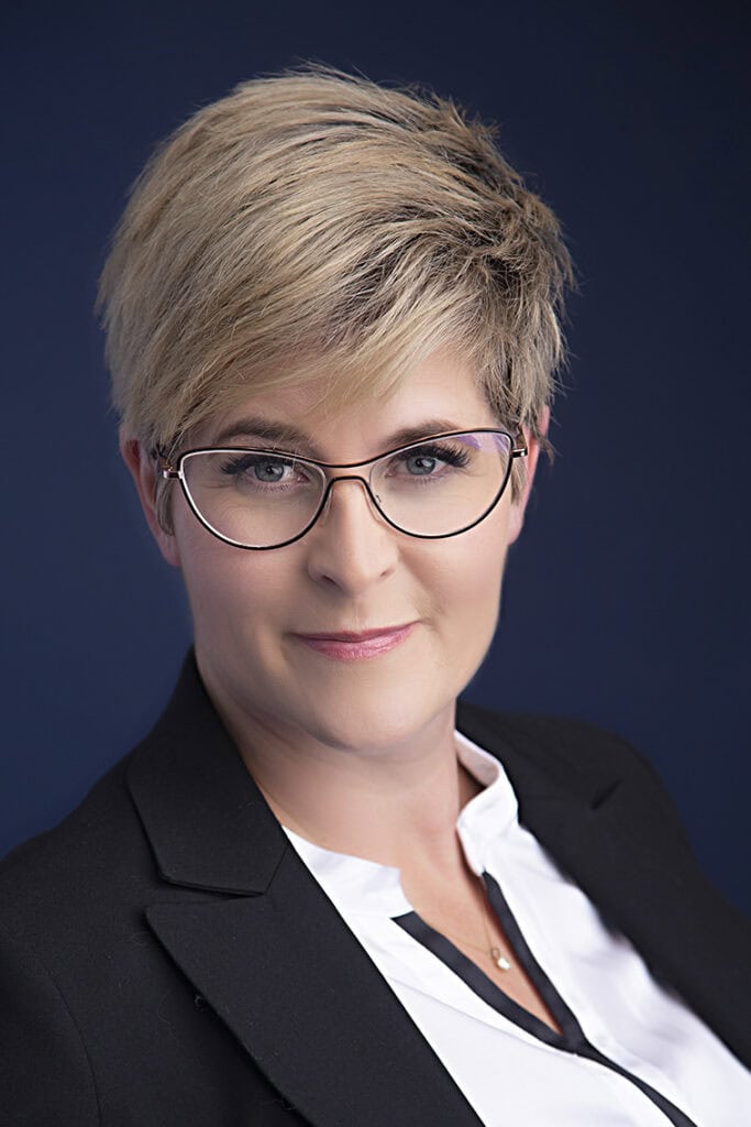 Professional woman with short blonde hair, wearing glasses, a black blazer, and a white blouse, exuding confidence and style as she looks directly at the camera against a dark blue background. - Jodi O Photography