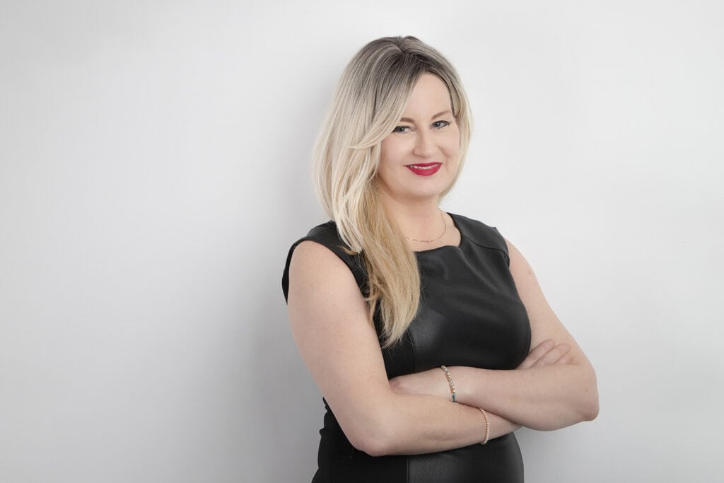 A woman with long, blonde hair wearing a black, sleeveless dress stands against a plain white background, smiling with her arms crossed—a powerful presence that exudes vibrant communication. - Jodi O Photography