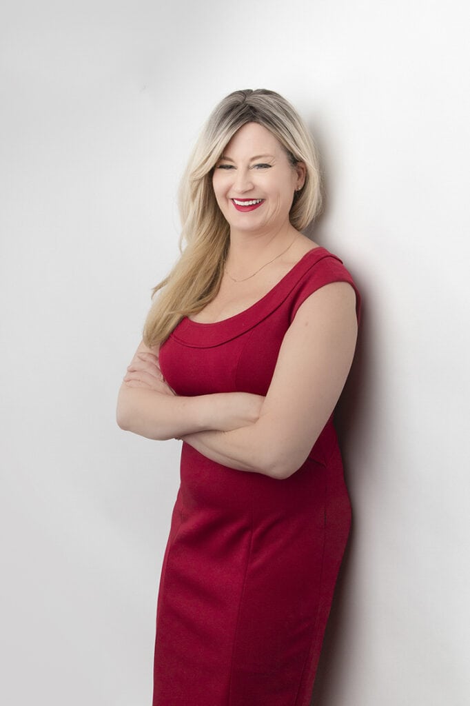 A woman with long blonde hair, wearing a sleeveless red dress, stands against a white background with her arms crossed, showcasing a powerful presence as she smiles at the camera. - Jodi O Photography