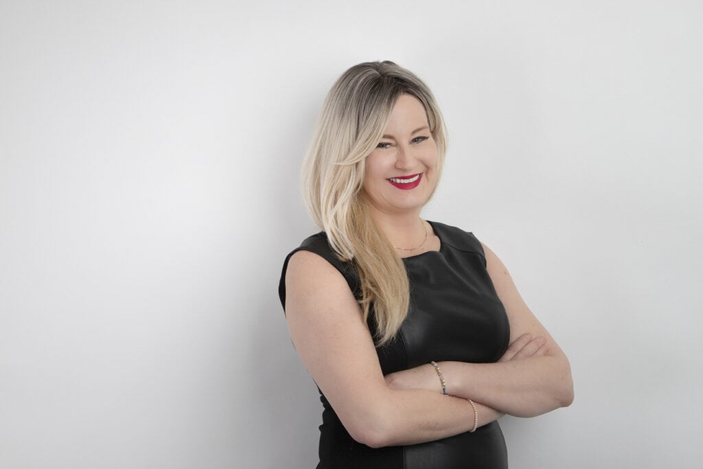 A woman with long blonde hair and red lipstick, wearing a sleeveless black top, stands against a plain white background with her arms crossed, exuding a powerful presence and smiling confidently. - Jodi O Photography