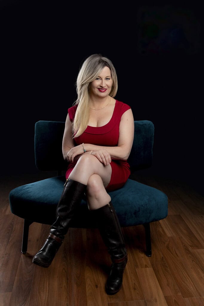 A woman with long blonde hair in a red dress and black boots sits on a dark teal chair, smiling with her legs crossed—her powerful presence radiates against the black background and wooden floor. - Jodi O Photography