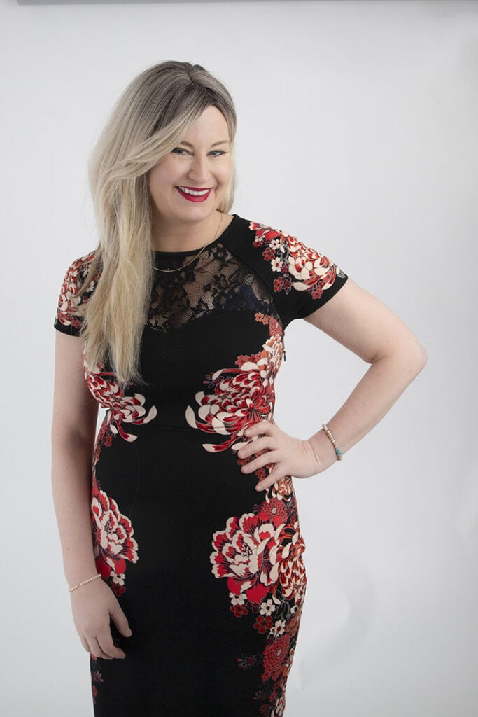 A smiling woman with long blonde hair and a powerful presence wears a black dress decorated with red and white floral patterns, standing against a plain white background with one hand on her hip. - Jodi O Photography